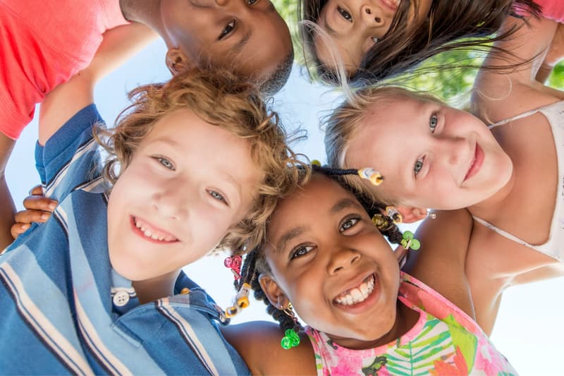 Fünf lachende Kinder stecken im Kreis die Köpfe zusammen und schauen von oben in die Kamera.